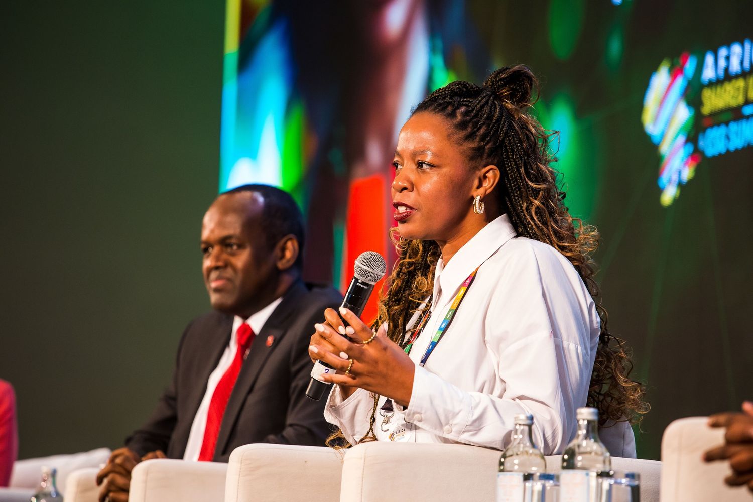 A speaker in a bright pink blazer presents from a podium on stage at a professional conference event.