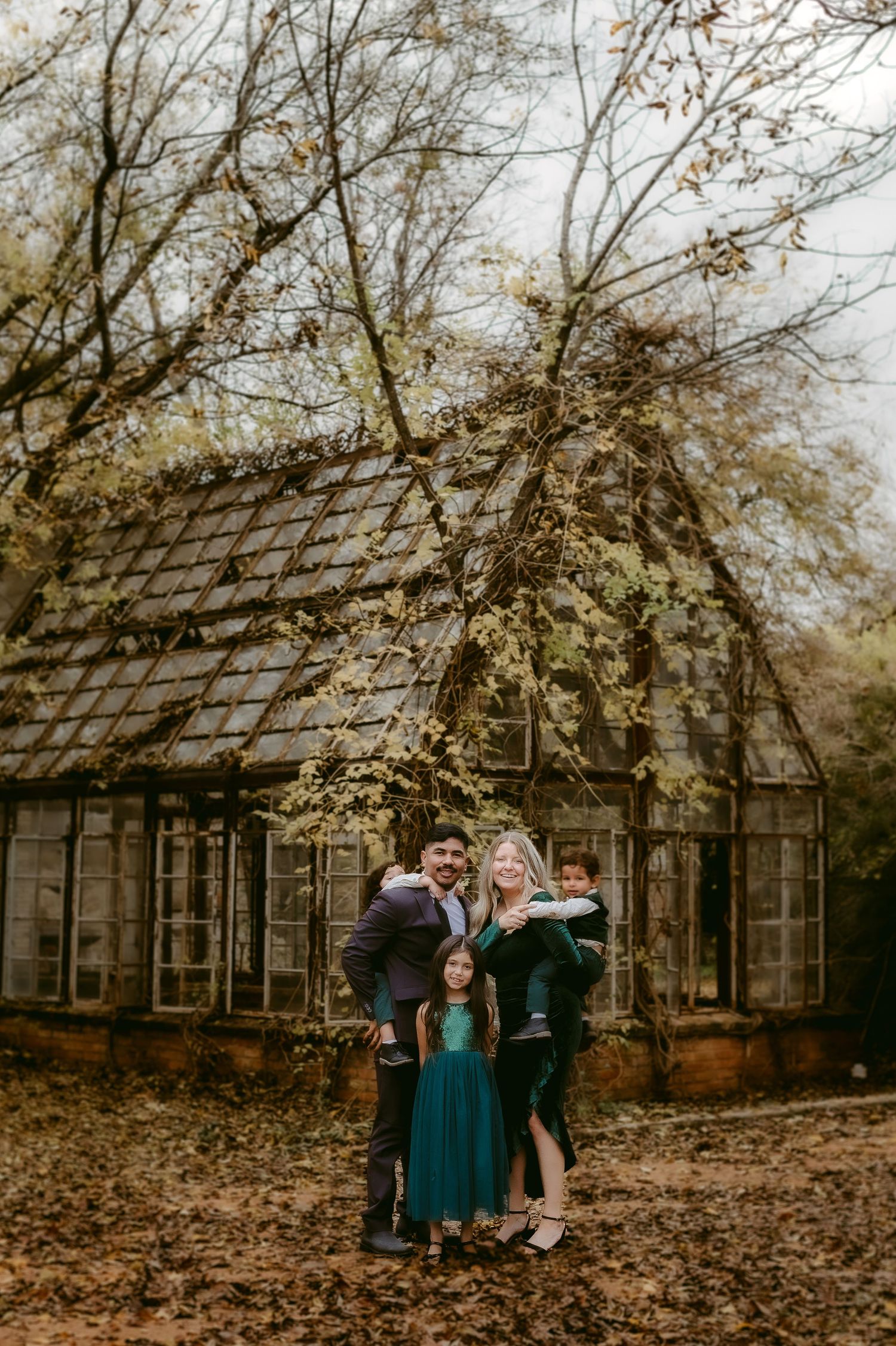 A rustic wooden greenhouse structure provides a charming backdrop for a fall family photoshoot among scattered leaves.