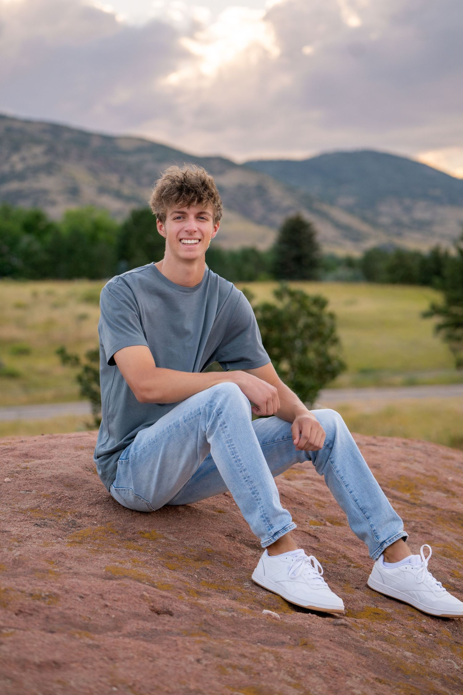 A person in jeans and a grey t-shirt sits casually on red rocks with mountains and green fields in the background.