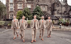 Groomsmen in beige suits line up playfully against historic brick building backdrop.