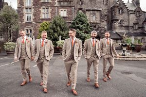 Groomsmen in tan suits strike fun poses while jumping in front of a historic stone building.