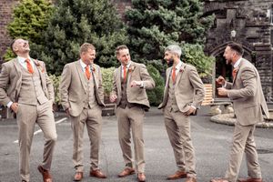 Groomsmen in tan suits pose playfully in a line outdoors with greenery in the background.