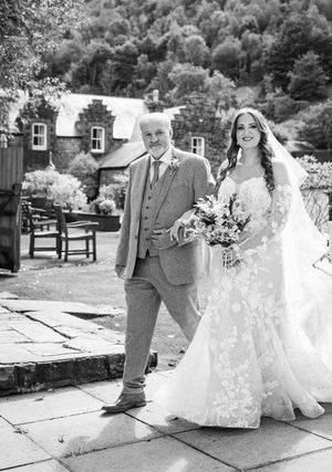 Elegant wedding couple poses along stone pathway with rustic buildings in background.