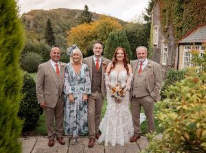 Wedding party poses outdoors in matching beige suits and floral accents against stone building and hills.