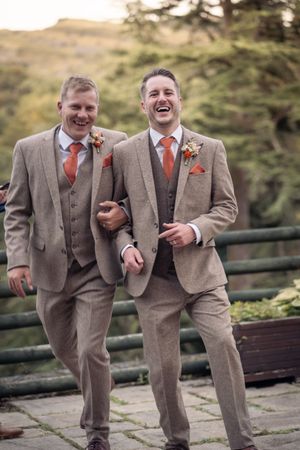 Two groomsmen in tan suits and coral ties share a laugh on garden terrace.