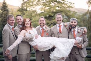 Bridal party wearing gray suits and coral ties celebrate joyfully together in an outdoor setting during golden hour.