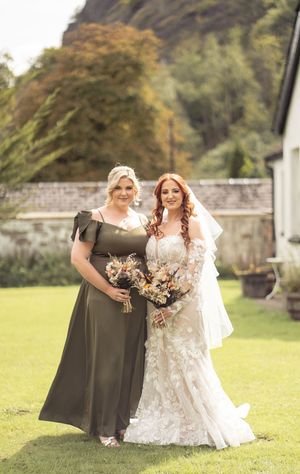 A formal bridal party poses on green grass beside stone walls with autumn foliage.