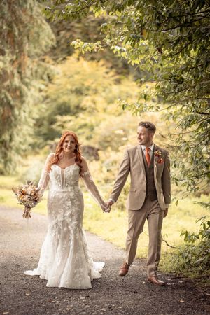 Couple holding hands while walking along a scenic wooded path during their wedding photoshoot.
