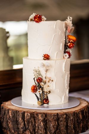 Two-tier white wedding cake decorated with orange flowers and dried botanicals on a rustic wood slice base.