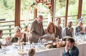 Wedding guests seated at long tables inside a bright reception venue with large windows and natural lighting.