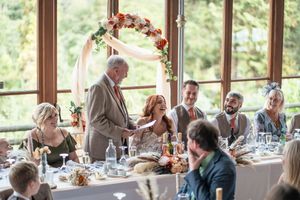 Wedding speech being given at a reception table decorated with floral arch and natural window lighting.