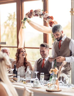 Wedding guests share joyful moments at a sunlit reception dinner table decorated with floral arrangements.