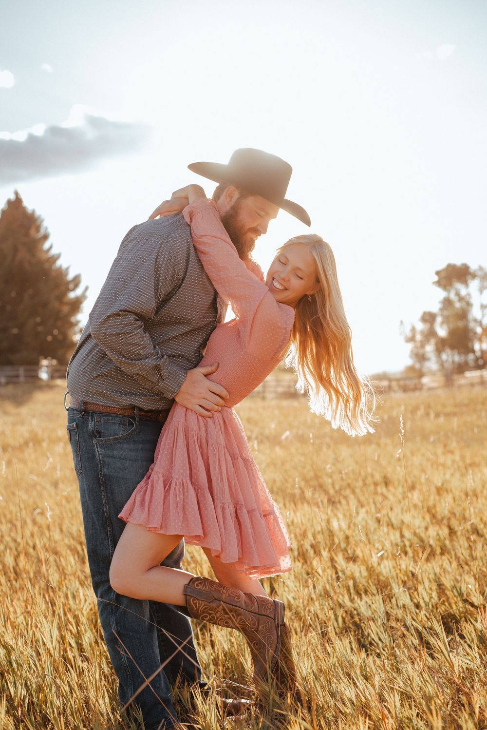 Romantic couple embraces in a golden wheat field at sunset with cowboy hat and pink dress.