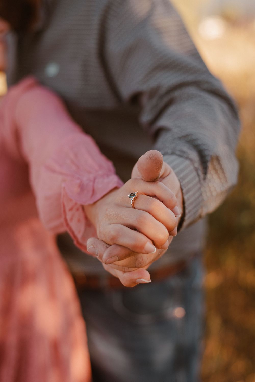 Close-up of hands holding with engagement ring in warm sunset light.