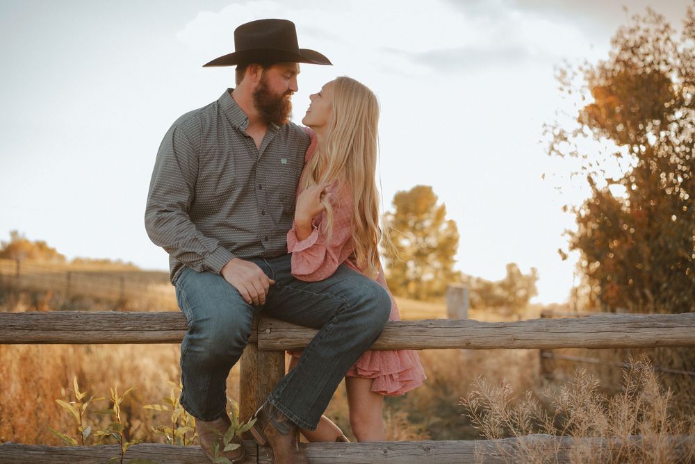 A romantic western-themed couple sits together on a wooden fence at sunset in an autumn field.