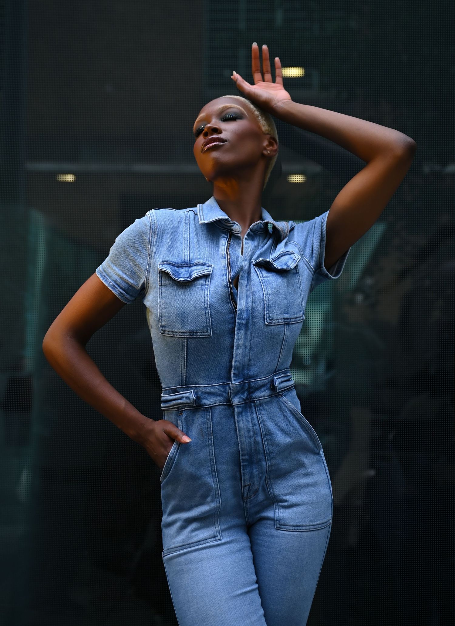 A person poses in a light blue denim jumpsuit against a dark background in a creative fashion photography series.