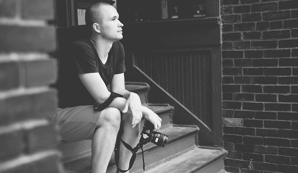 Black and white photo of young man sitting on steps outside brick building holding a camera strap.