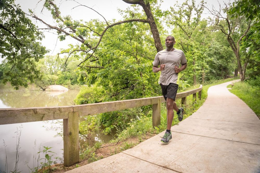 Male runner jogging on a scenic park trail beside a wooden fence and green foliage near water.