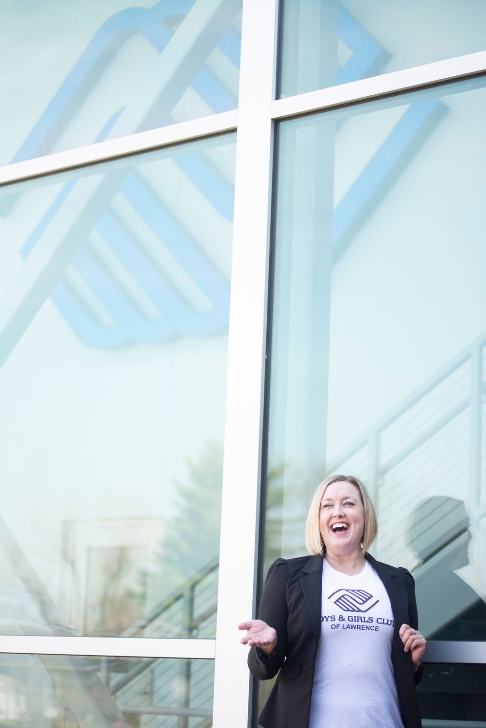Laughing blonde woman in Boys and Girls Club t-shirt and blazer posing by a large glass window.