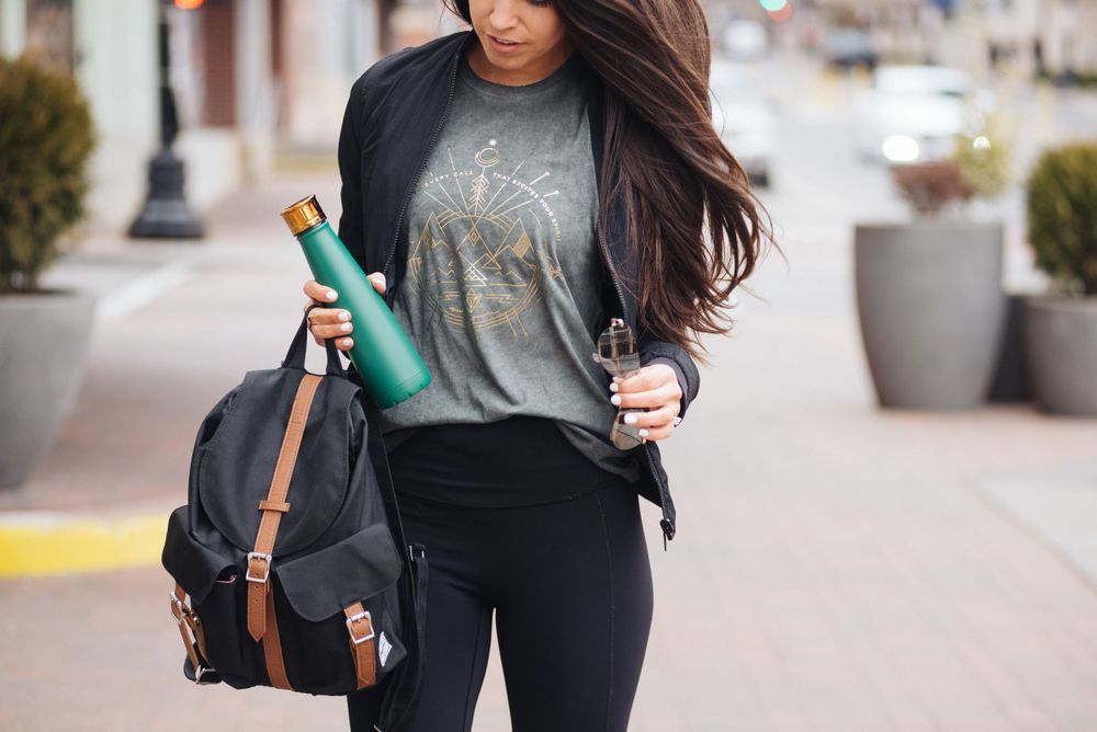 Woman with dark hair carrying a backpack and green water bottle walking down a brick sidewalk.