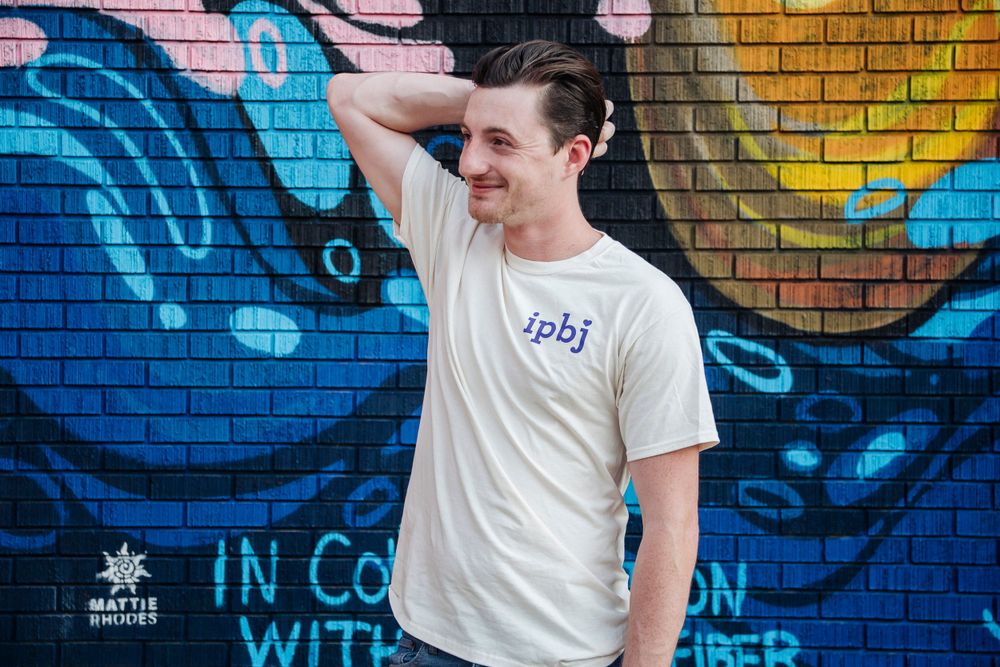 Young man in white IPBJ t-shirt smiling in front of colorful urban street art mural.