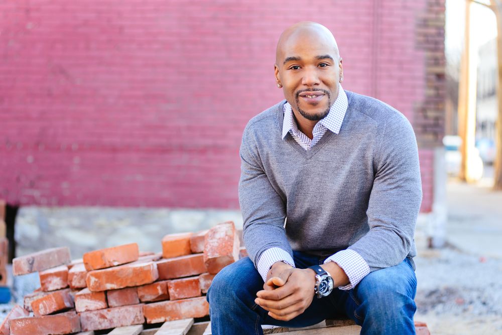 Smiling man in a grey sweater sitting on bricks in front of a red brick wall.