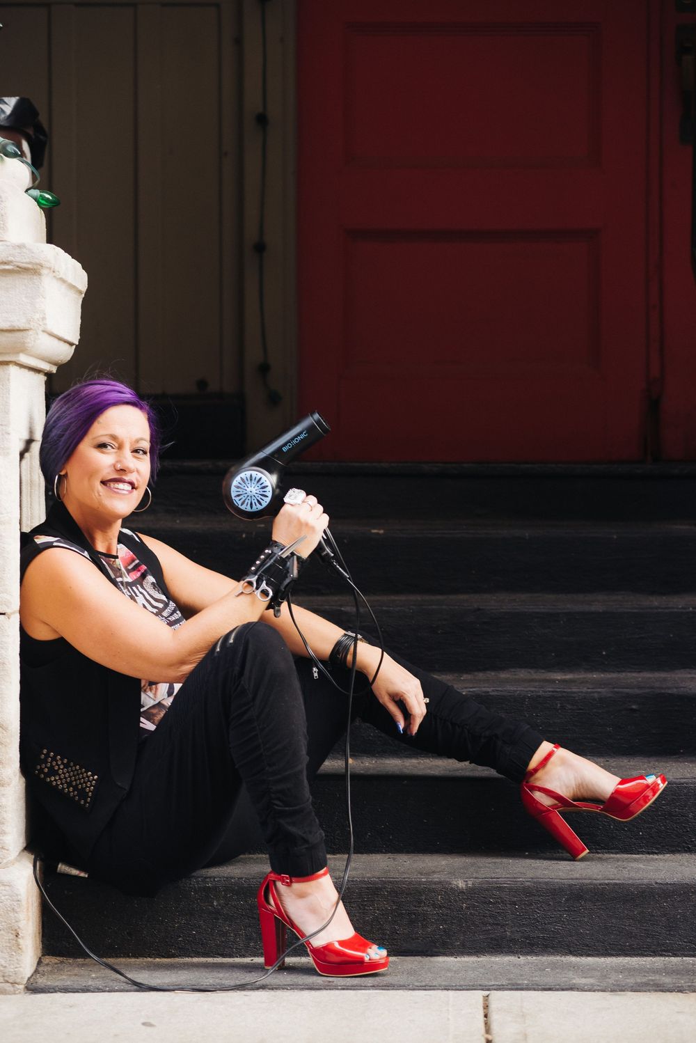 Woman with purple hair sits on steps holding a microphone wearing red heels and black outfit.