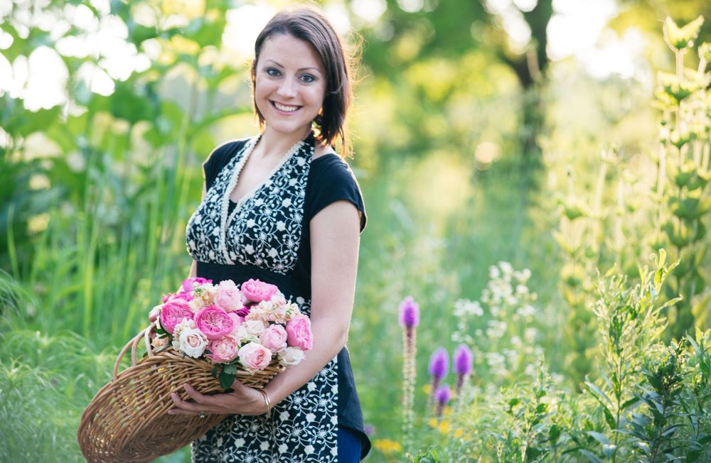 A smiling woman holds a wicker basket of pink and white roses in a lush green garden.