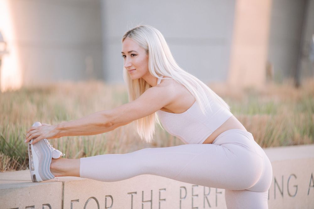 Blonde woman in white athletic wear stretching outdoors near a concrete structure.