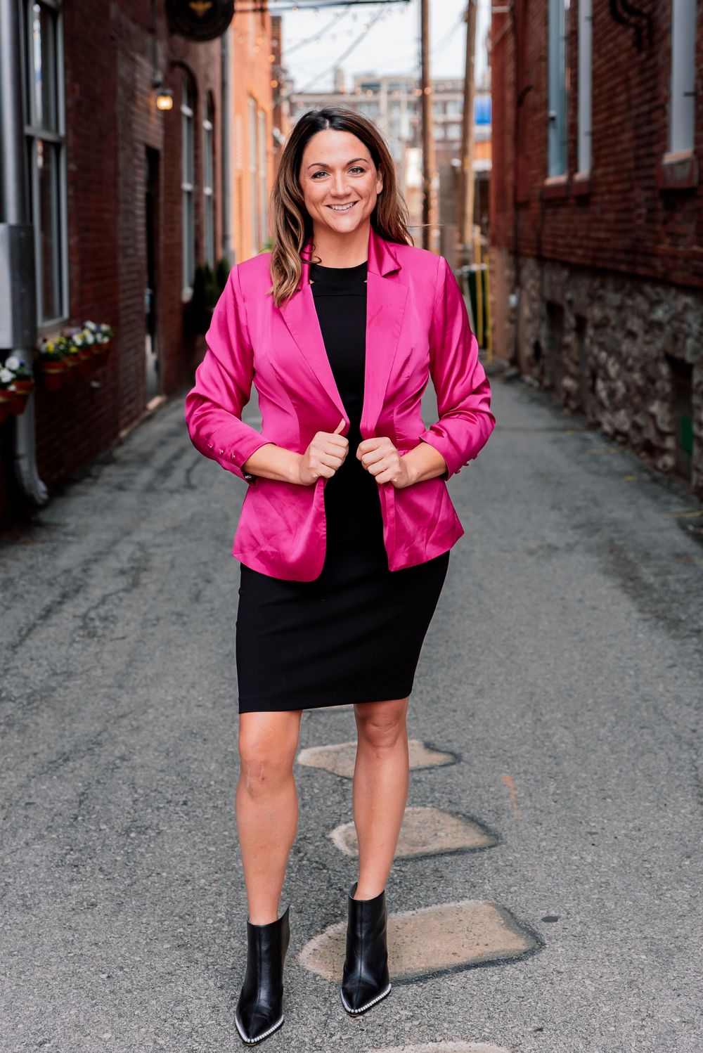 Woman in pink blazer and black dress smiles confidently while standing in a brick alleyway.
