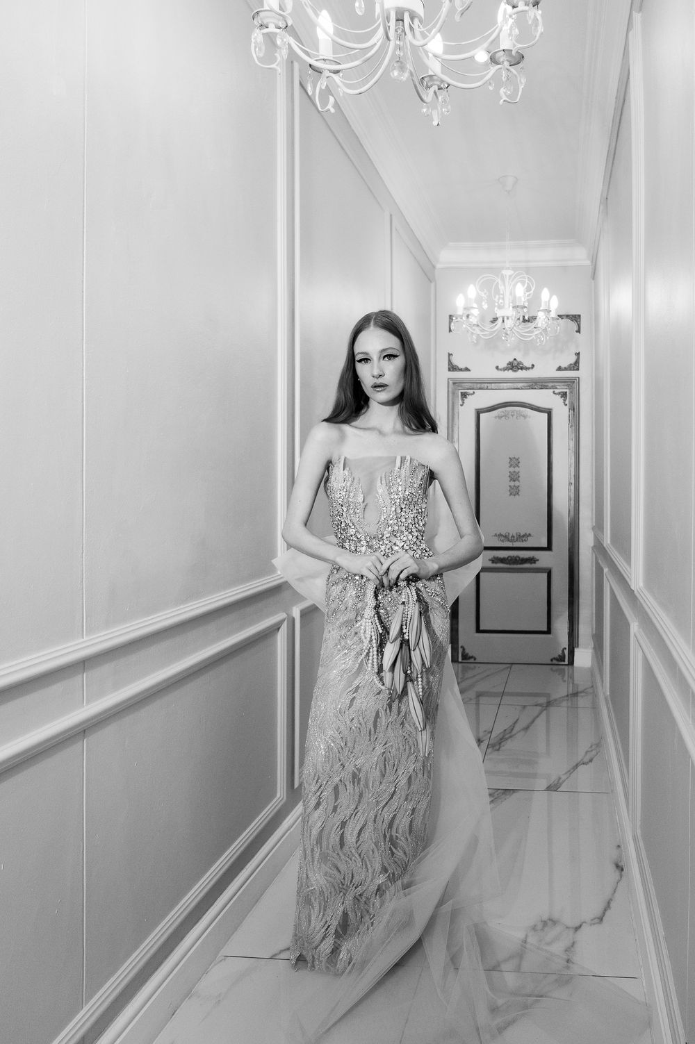 Black and white photo of elegant gown in ornate hallway with chandeliers and marble floors.