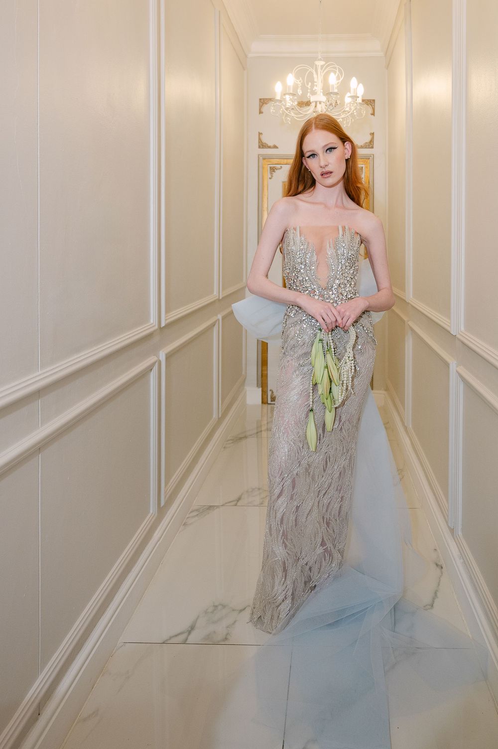 A woman in an elegant blue and silver sequined gown walks through a luxurious white hallway with a crystal chandelier.