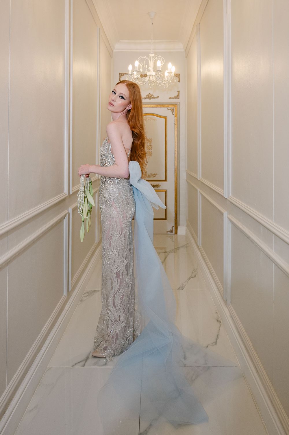 A dramatic evening gown with blue tulle flows through an elegant white corridor with wall moldings.