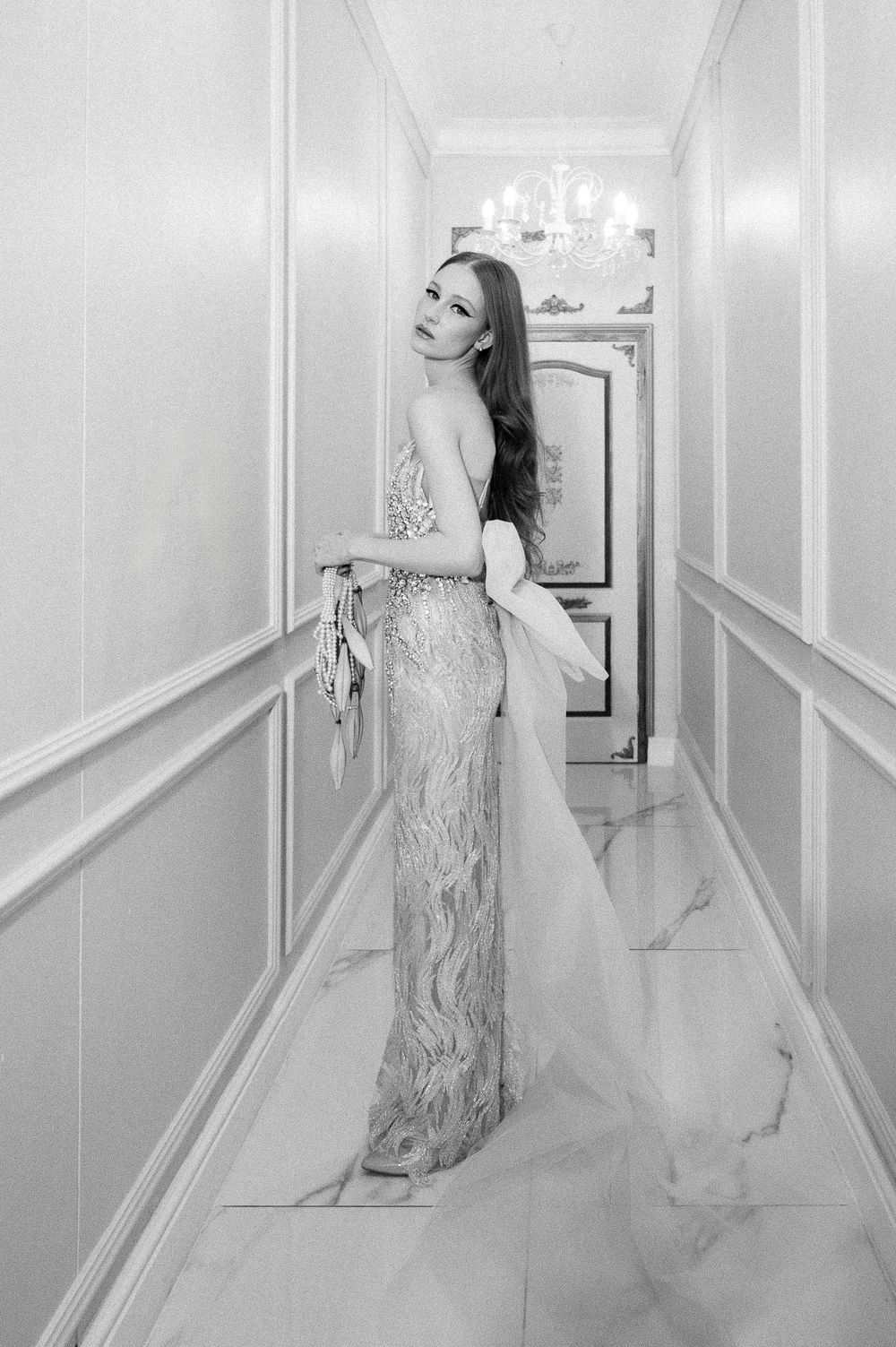 An elegant figure poses in a flowing silver gown against white paneled hallway walls with a crystal chandelier.
