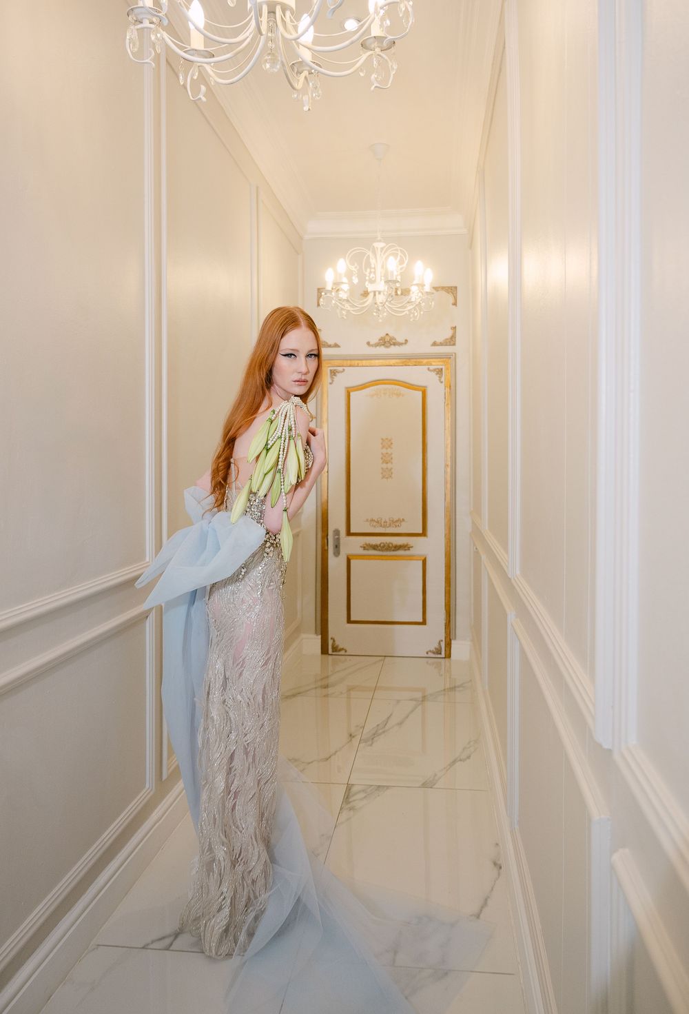 Elegant figure in a flowing gown poses gracefully in a bright white hallway with ornate wall moldings and chandeliers.