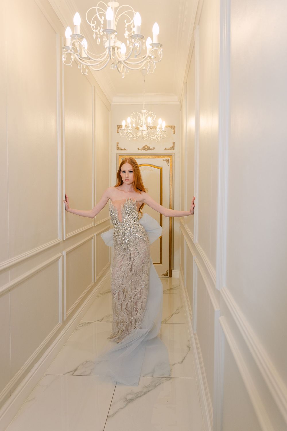 Figure in shimmering gown moves gracefully through elegant white hallway with chandelier lighting.