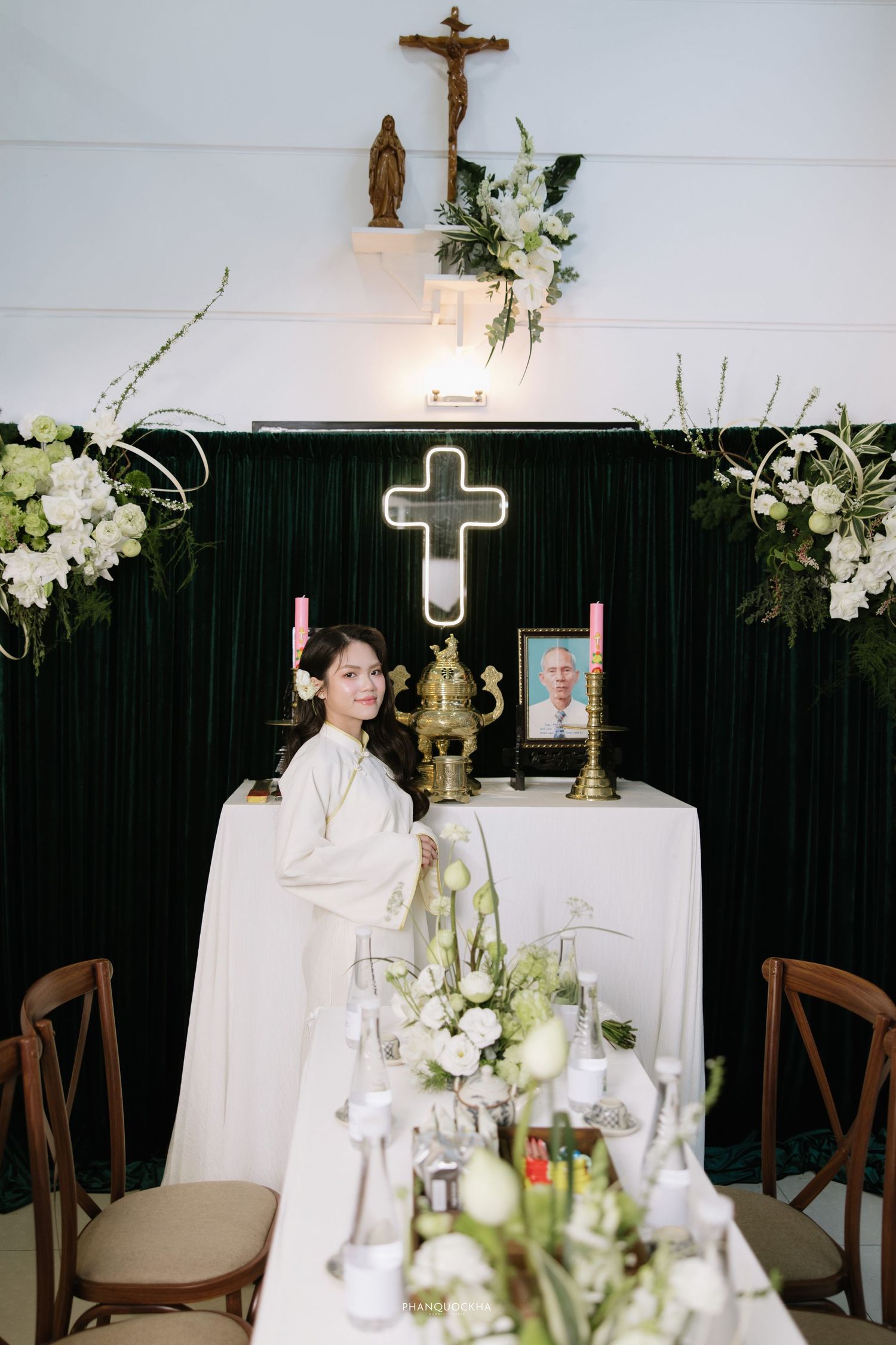 Young woman in white ao dai standing before a Catholic altar with cross, candles, and white flowers.