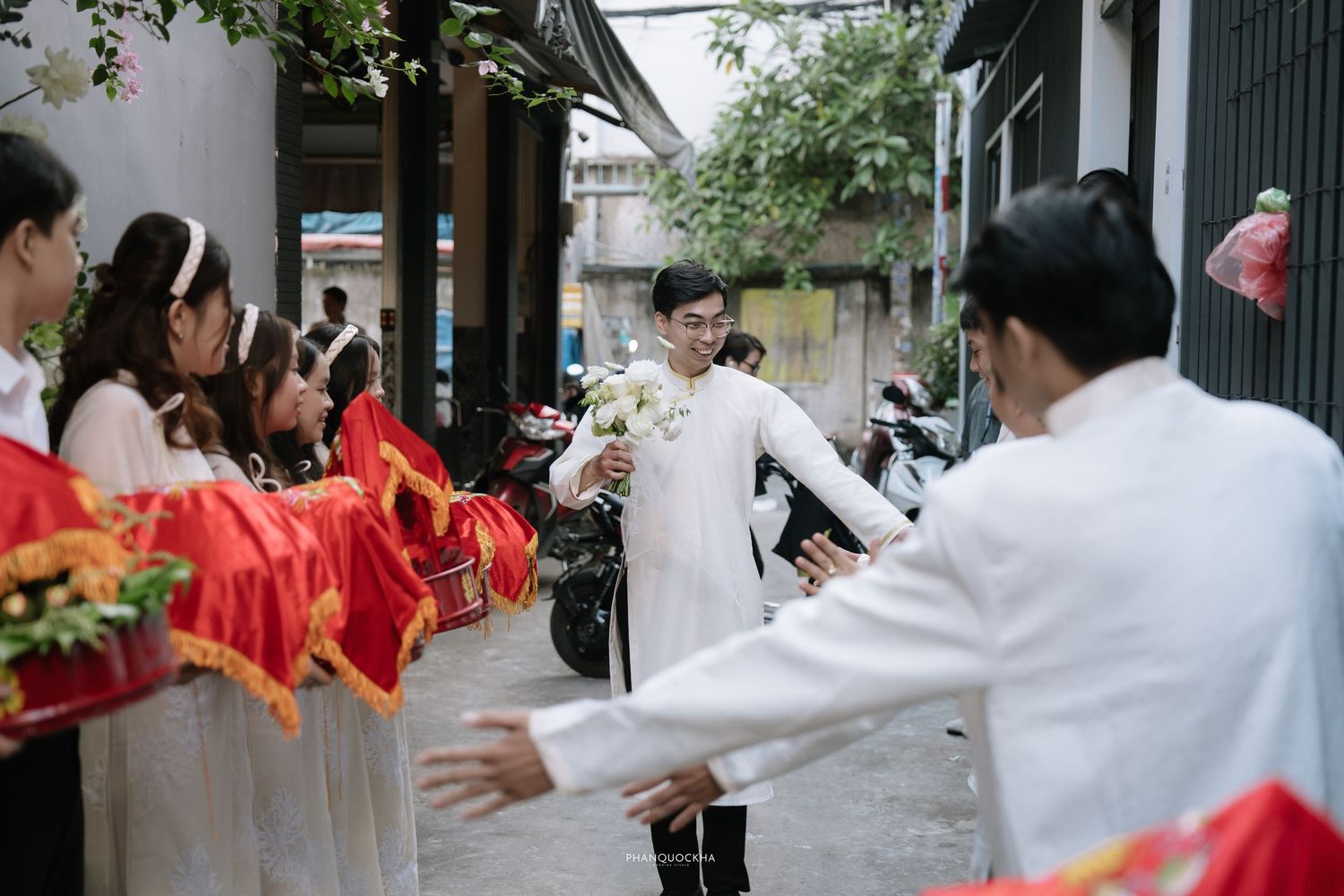 Groom in white ao dai arriving with wedding party carrying red ceremonial gifts at a traditional Vietnamese wedding.