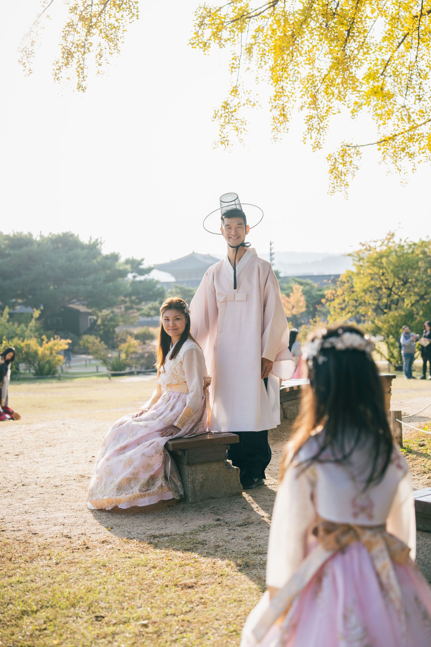 A couple in traditional Korean hanbok attire poses together in a park during golden hour.