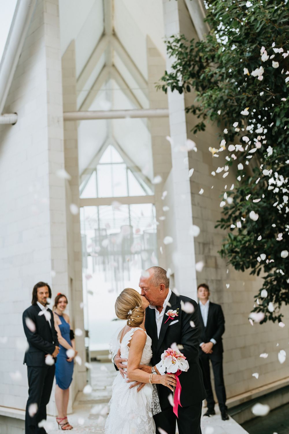 Celebratory moment with flower petals falling in front of white architectural backdrop.