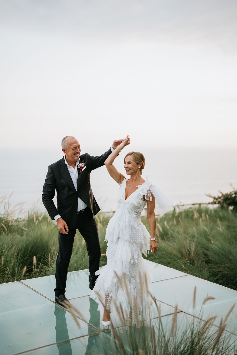 Couple dancing on reflective platform surrounded by coastal grass at outdoor venue.