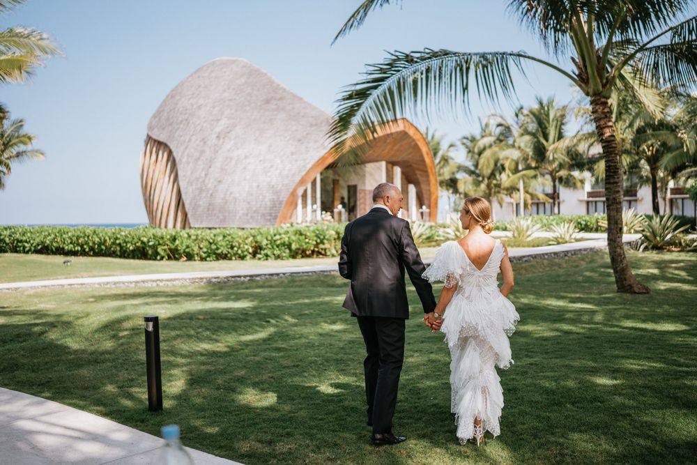 Wedding couple walking on lawn path near modern architectural structure with palm trees.