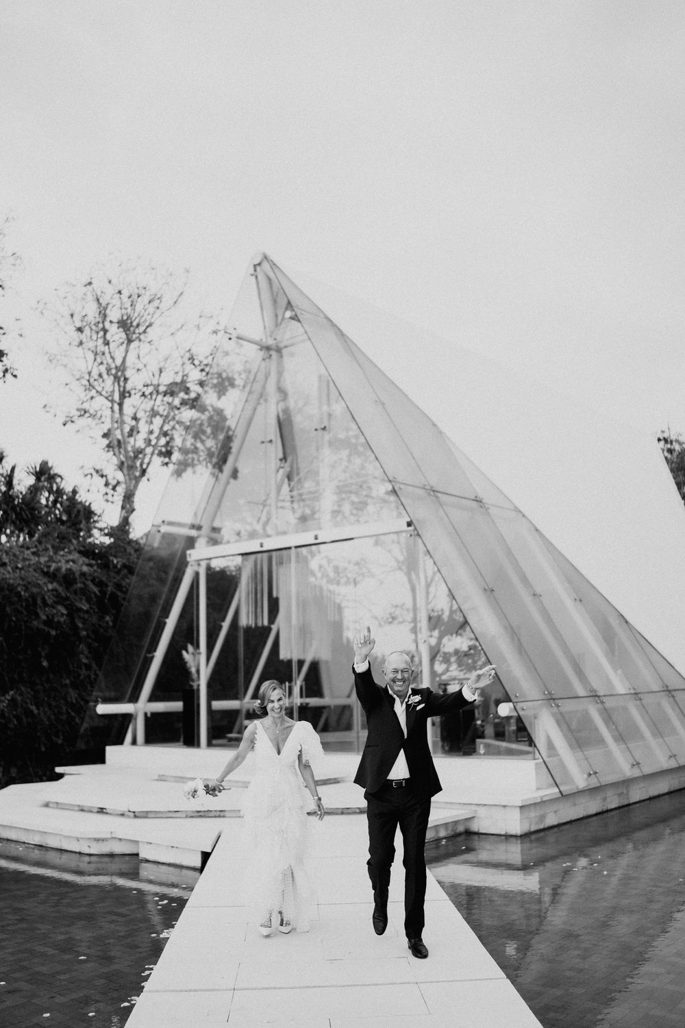 Wedding couple walking near modern glass pyramid structure in black and white photography.