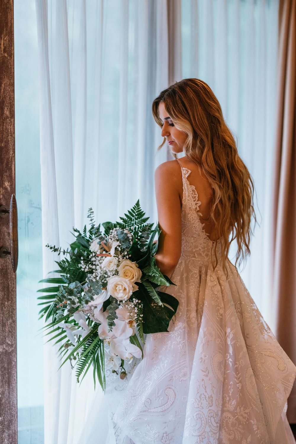 An elegant lace wedding dress with a bouquet of white roses and greenery against white curtains.