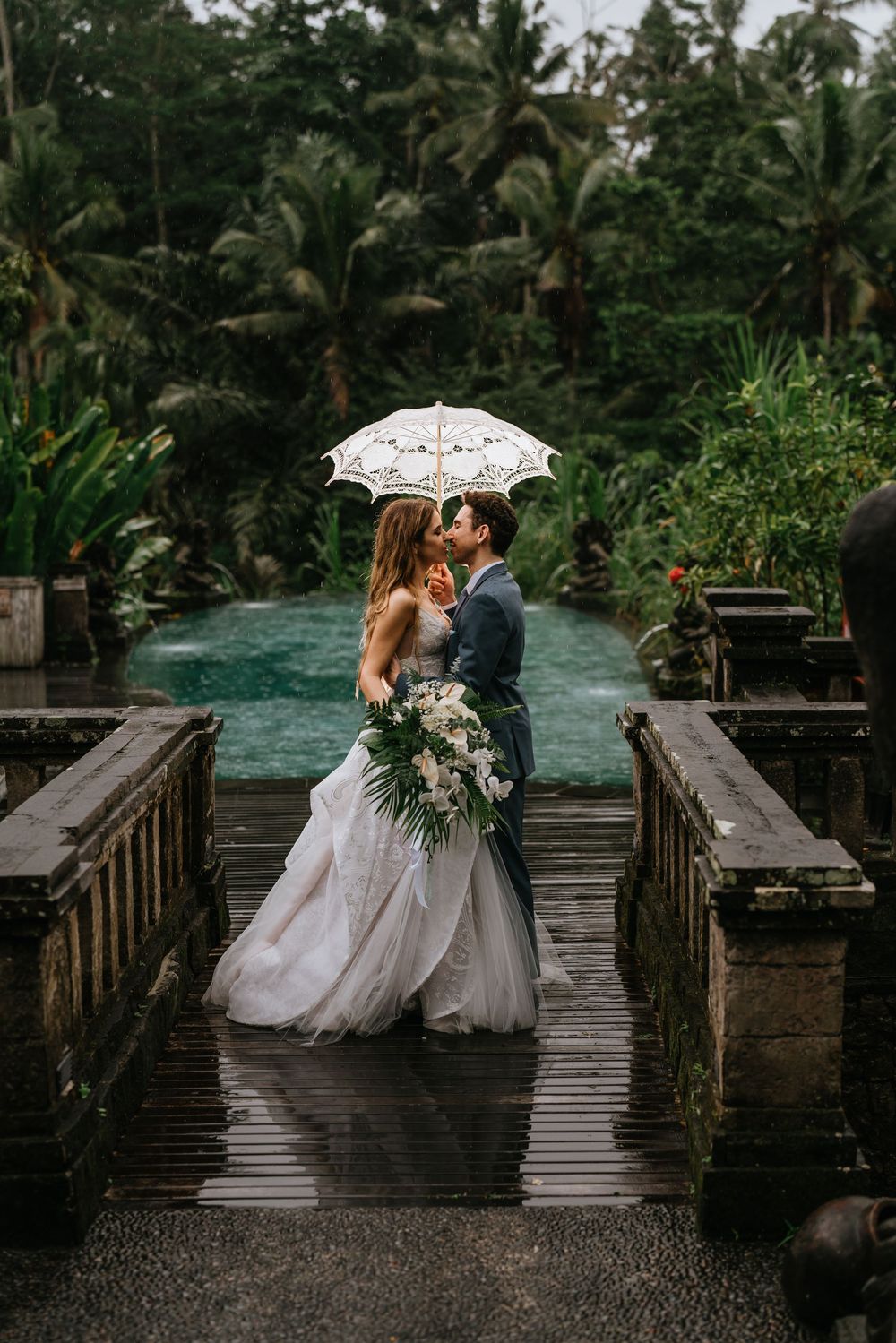 Wedding couple shares intimate moment on wooden boardwalk surrounded by tropical rainforest in the rain.