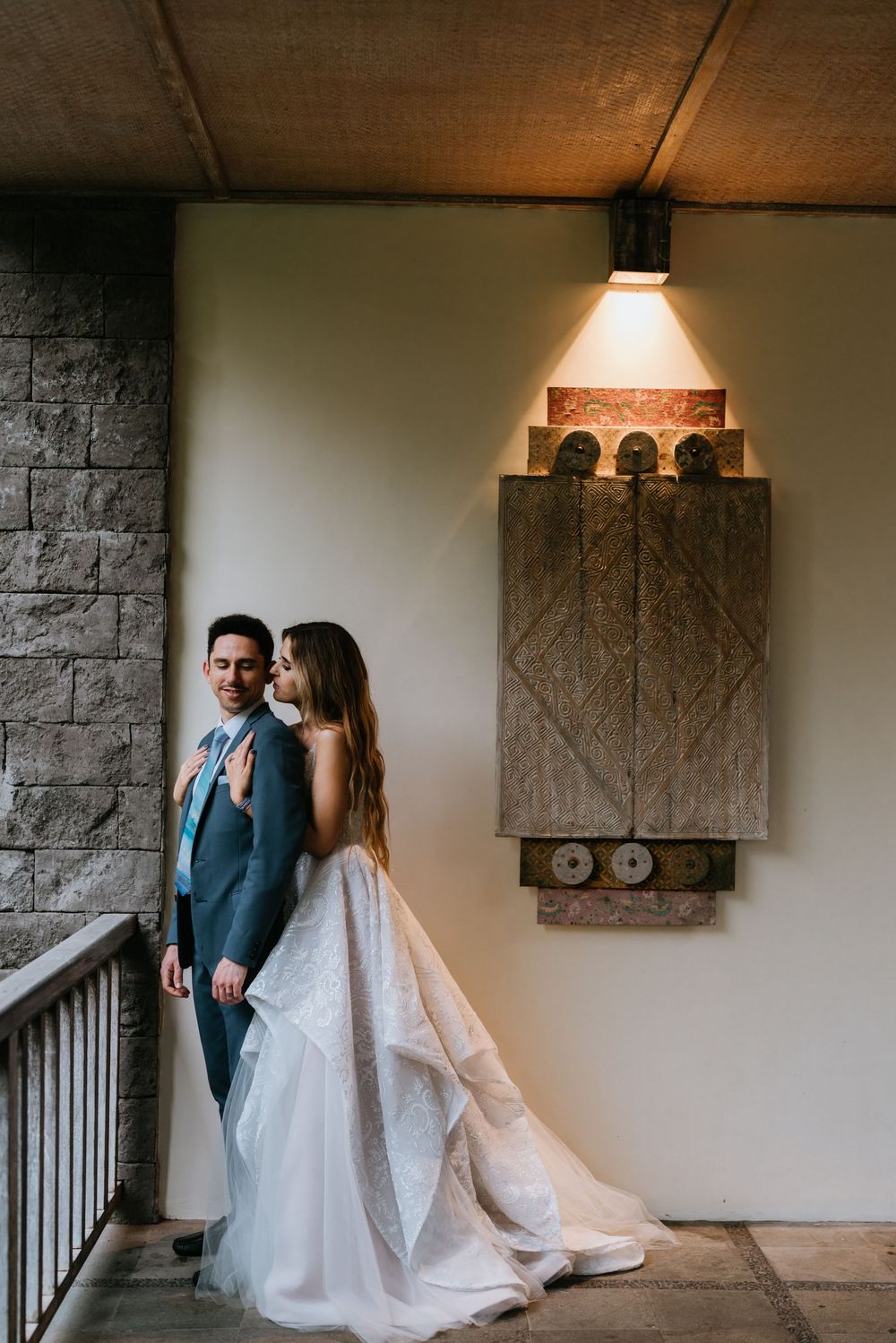 Elegant wedding couple poses against stone wall interior with dramatic lighting and architecture.