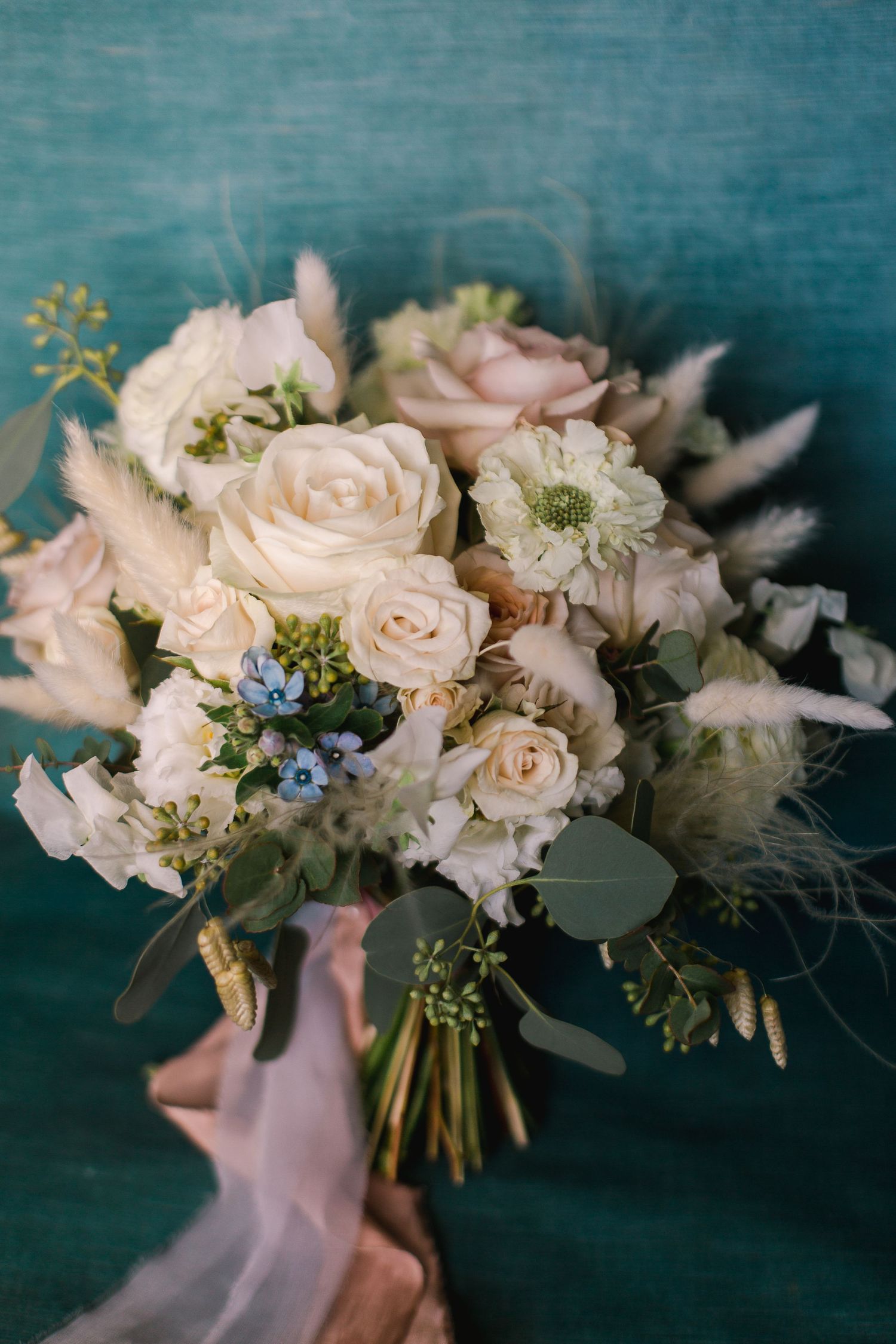 Elegant bridal bouquet with blush roses, blue accents and delicate greenery against a teal background.
