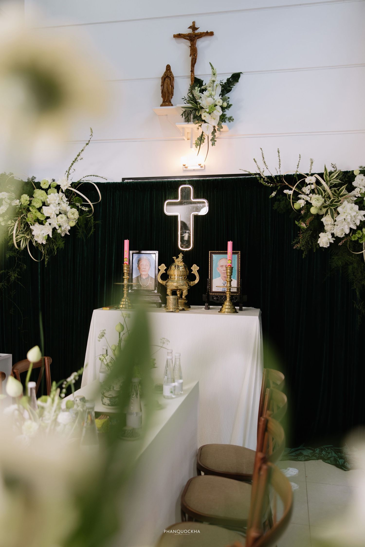 Christian wedding altar adorned with white flowers, candles, cross, and framed photos on a draped table.