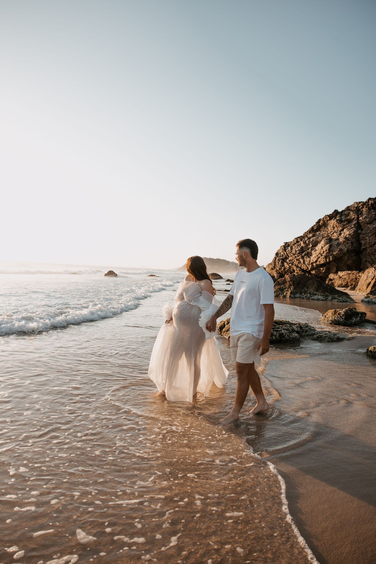 Ethereal beach photoshoot with flowing white dresses against ocean backdrop.