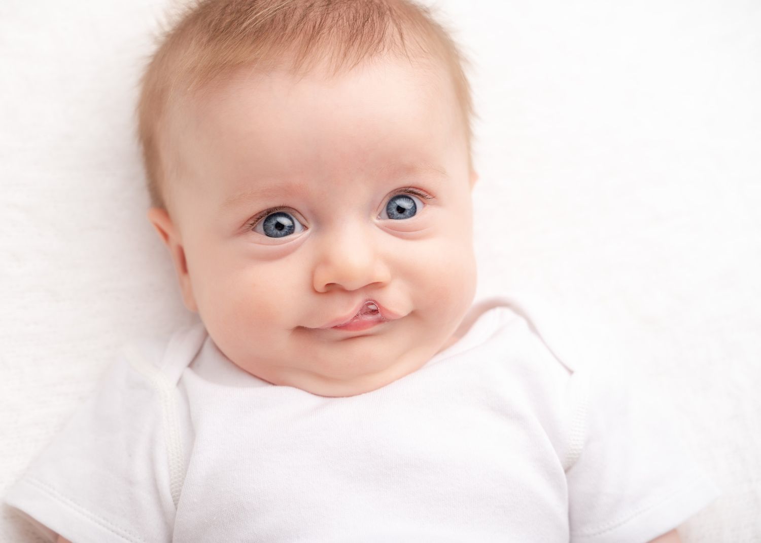 Close up portrait of a baby with bright blue eyes wearing white against a white background.